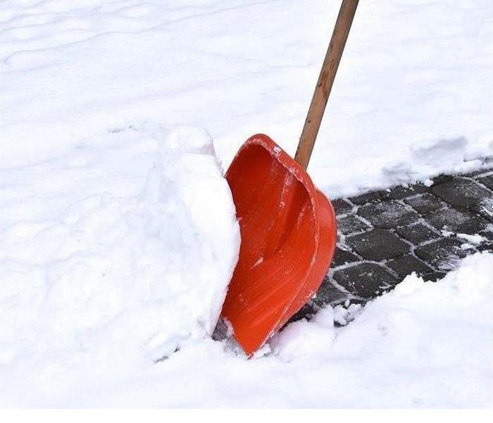 The lower part of a snow shovel moving a scoop of snow from a snow-covered sidewalk that is brick paved